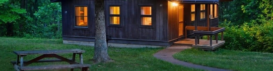 Cabin exterior illuminated at dusk with a fire ring in the foreground and trees surrounding the property.