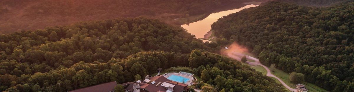 Aerial view of Shawnee Lodge surrounded by rolling forested hills at sunset, with a winding river visible in the distance and warm evening light illuminating the landscape.